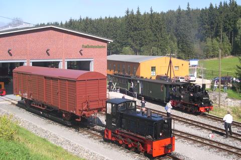 99 4511 hält gerade am Bahnübergang an der Ausstellungs- und Fahrzeughalle, während die Ns4 199 007 der Preßnitztalbahn mit dem aufgebockten Hbs vor dem Tor 3 der Halle steht.